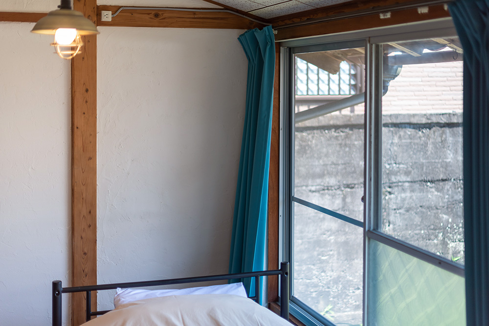 写真：窓際に置かれたベッド。カーテンはターコイズブルー