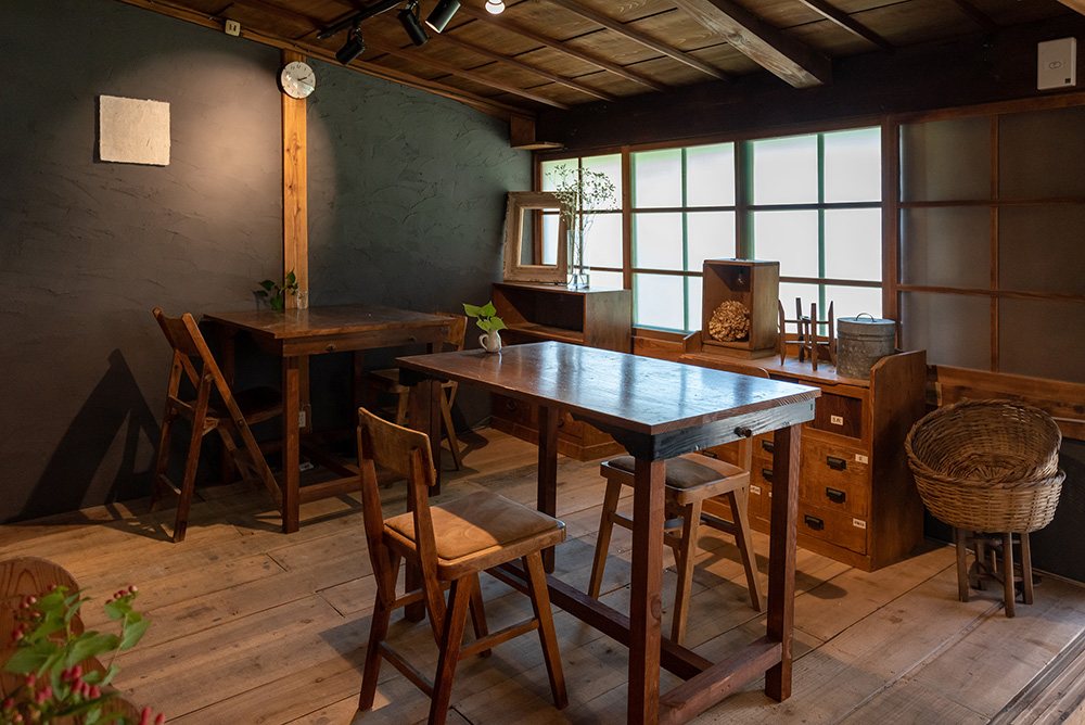 写真：2人用のテーブルが2つ並んだカフェスペース。床は板張り、壁はシックなグレー。窓際にはアンティークな箪笥や雑貨が並んでいる。