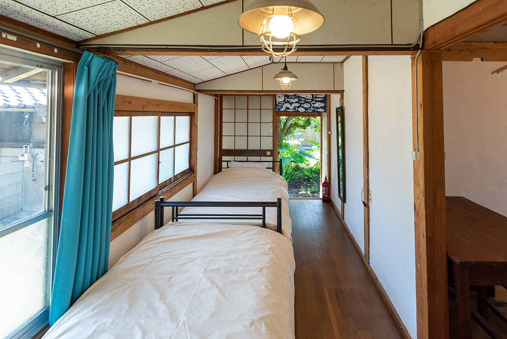 写真：2つの部屋がつながった客室のはなれ。洋間には2つのベッドが縦に並んでいる。カーテンはターコイズブルー。玄関の外に庭の緑が見える。