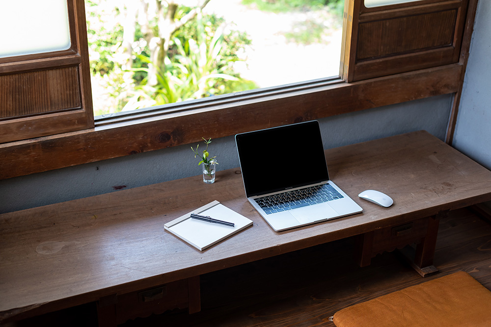写真：窓際の文机。机の上には mac book とマウス、メモとペン、小さな一輪挿し。窓の外には庭の緑が見える。